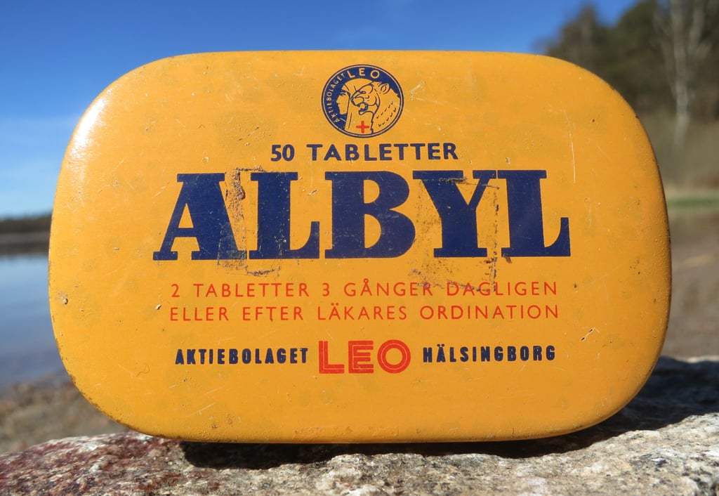 Gul, avlang blikkeske merket Albyl og 50 tabletter, liggende på en stein med blå himmel bak
