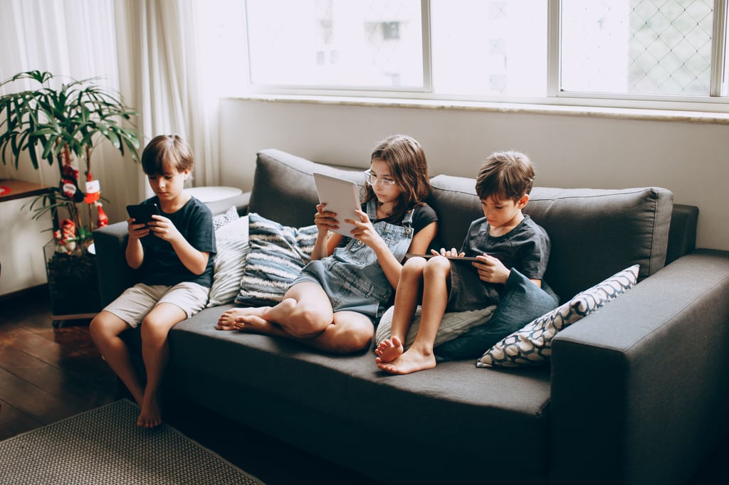 Tre barn sitter i en grå sofa og ser ned på hver sin mobil og nettbrett
