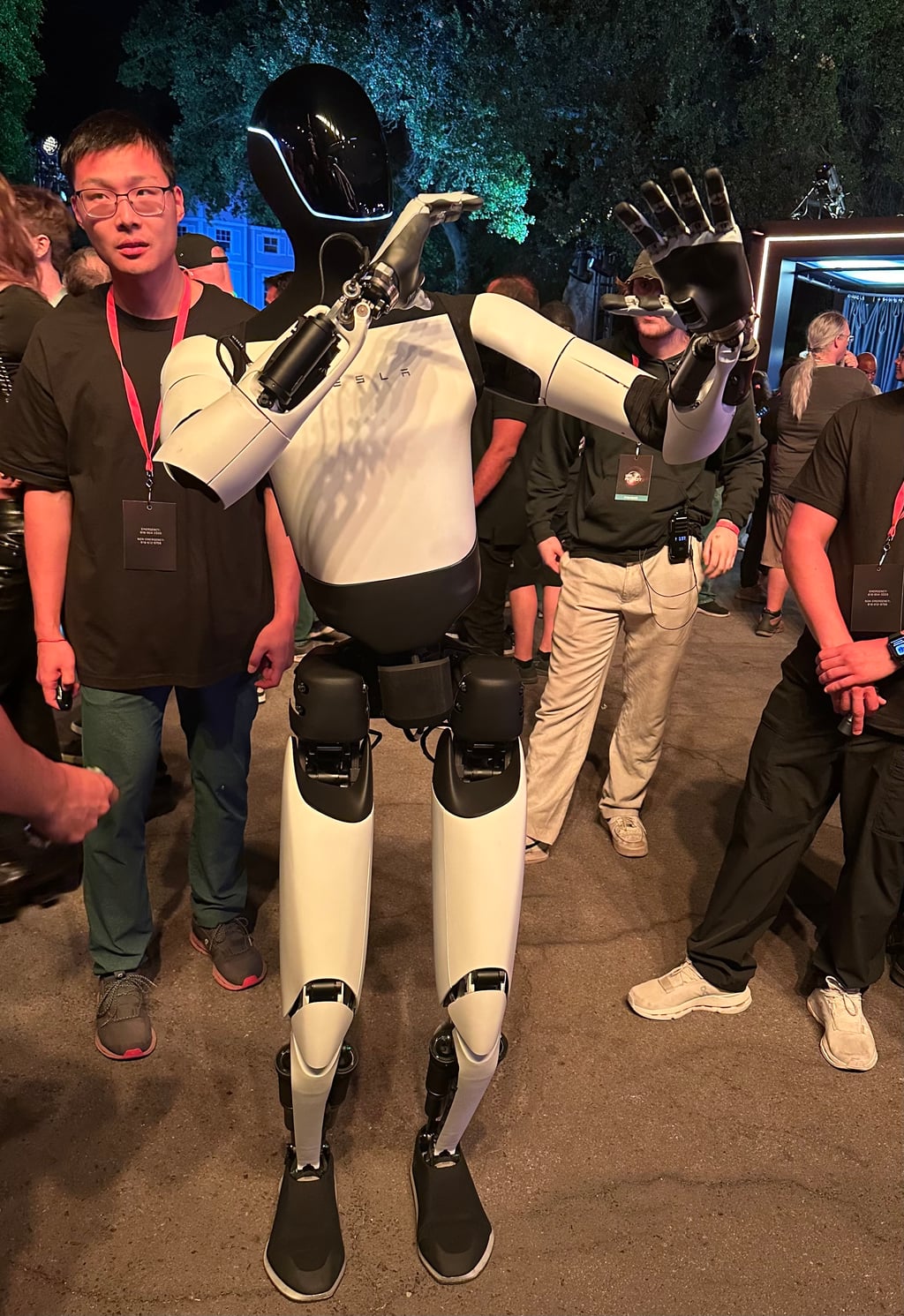 Hvit Tesla Optimus-robot med sort hode vinker med hånden mellom besøkende på et kveldsarrangement