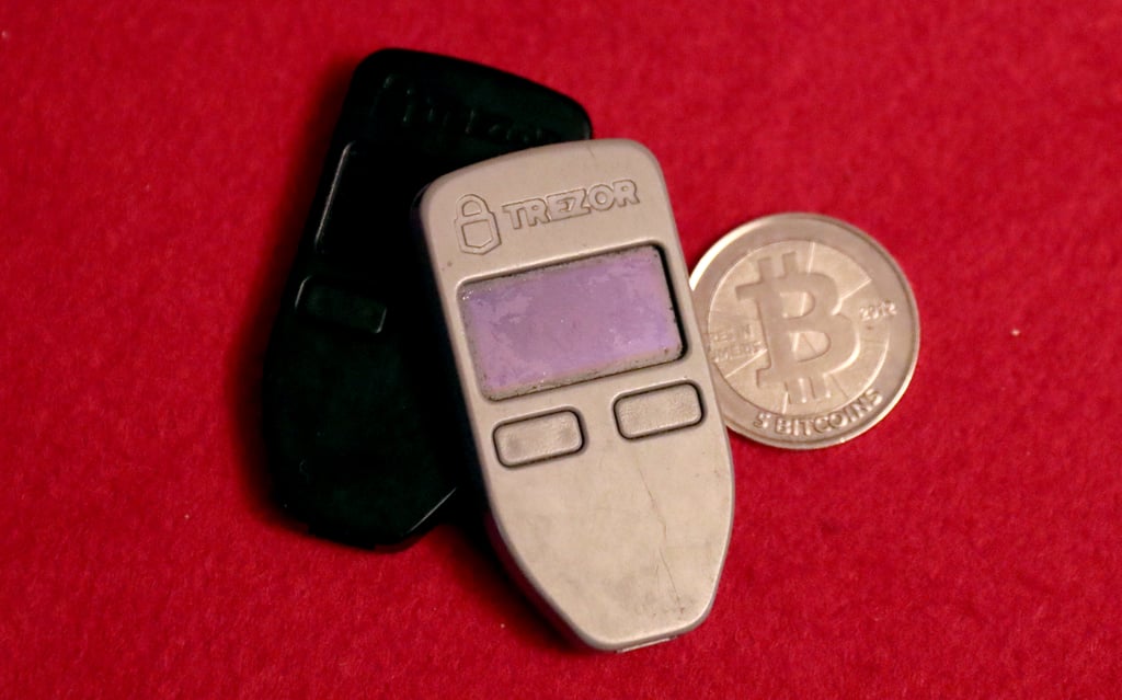 To Trezor-maskinvarelommebøker ved siden av en fysisk bitcoin-mynt på rødt underlag