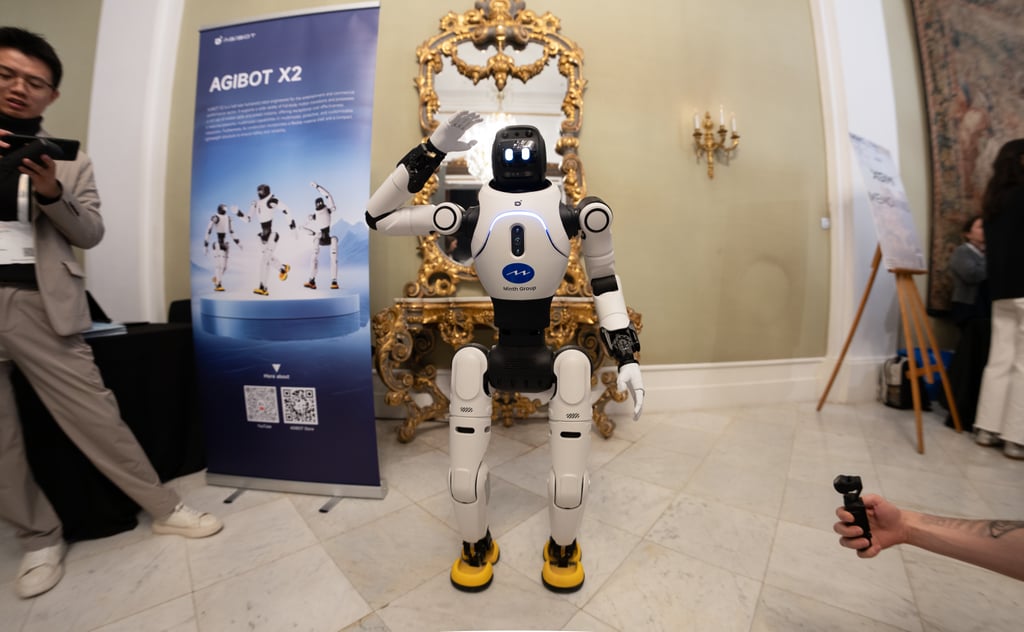 Hvit humanoid robot fra AgiBot står med hevet hånd foran en messevegg