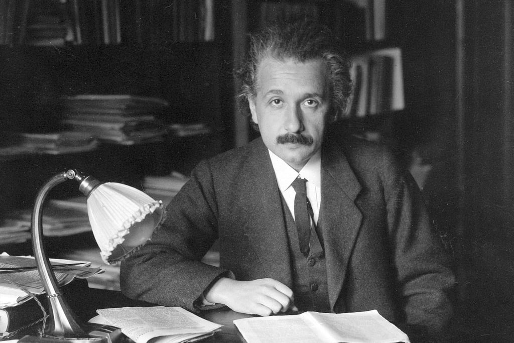 Albert Einstein sitter ved skrivebordet sitt med papirer og lampe, fotografert rundt 1920