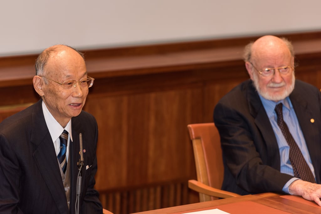 Satoshi Ōmura snakker i en mikrofon ved et bord i et rom med mørk trepanel. Til høyre sitter William C. Campbell med hvitt skjegg og briller og hører på.