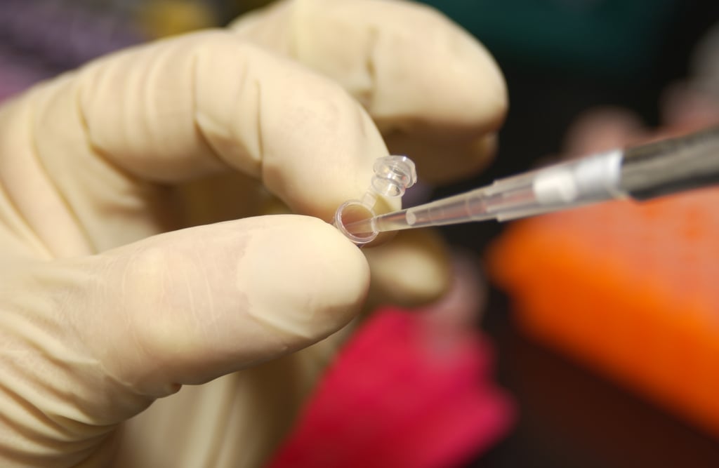 Hånd med lys latekshanske som holder et lite prøverør mens en pipette føres inn i det