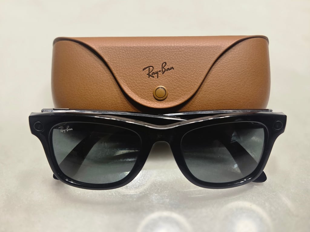 Sorte Ray-Ban Meta-smartbriller foran et brunt skinnetui, med kameralinsen synlig i innfatningen