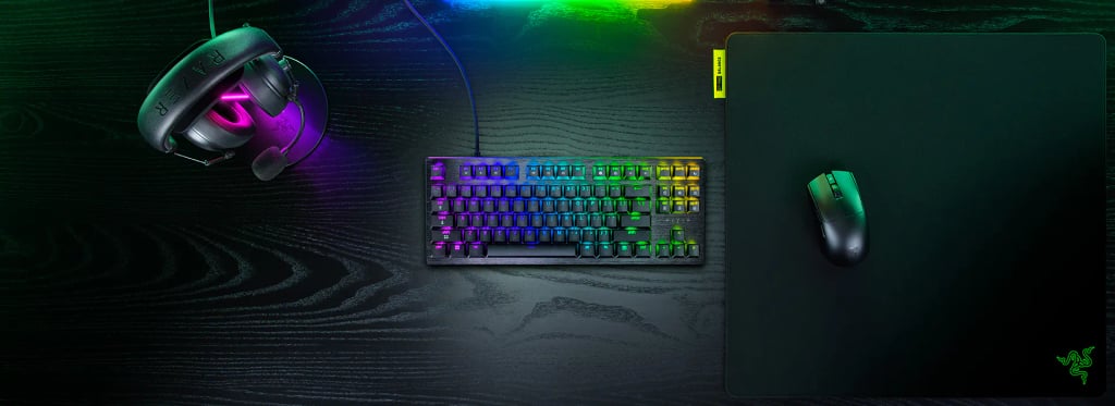 Et Razer-tastatur uten talltastatur med RGB-belysning på en mørk pult, med headset og mus ved siden av