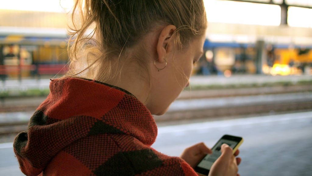 Ung kvinne i rødrutet jakke ser på mobiltelefonen sin mens hun venter på en togperrong