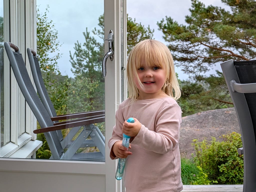 Et lite barn med lyst hår smiler mot kamera ved en åpen dør på en terrasse