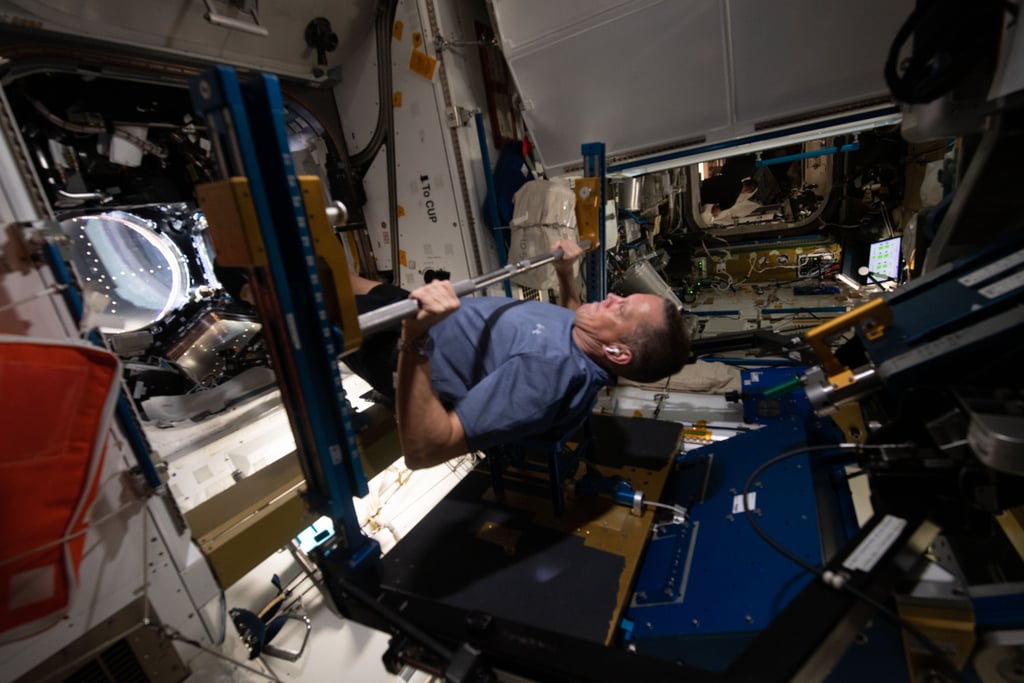 Astronaut Bob Hines løfter en stang i treningsmaskinen på romstasjonen, omgitt av utstyr og kabler