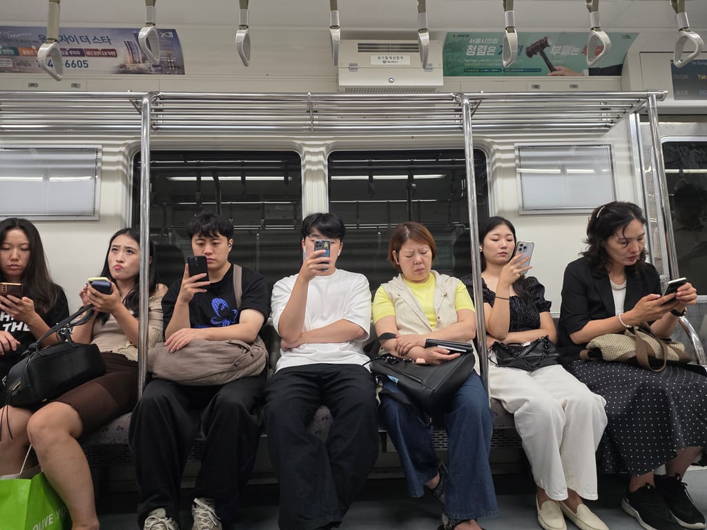 Syv passasjerer sitter på rekke i en T-banevogn i Seoul, de fleste med blikket i mobilen