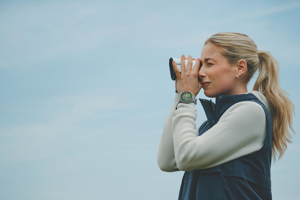 Kvinne i lys genser og blå vest ser gjennom en avstandsmåler, med Garmin Approach S72 på håndleddet