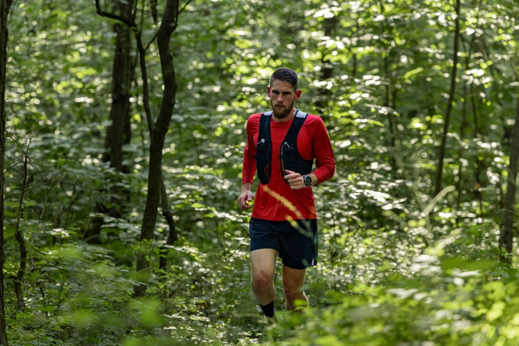 Løper i rød trøye og sort løpevest på vei gjennom løvskog, med Garmin Enduro 4 på håndleddet