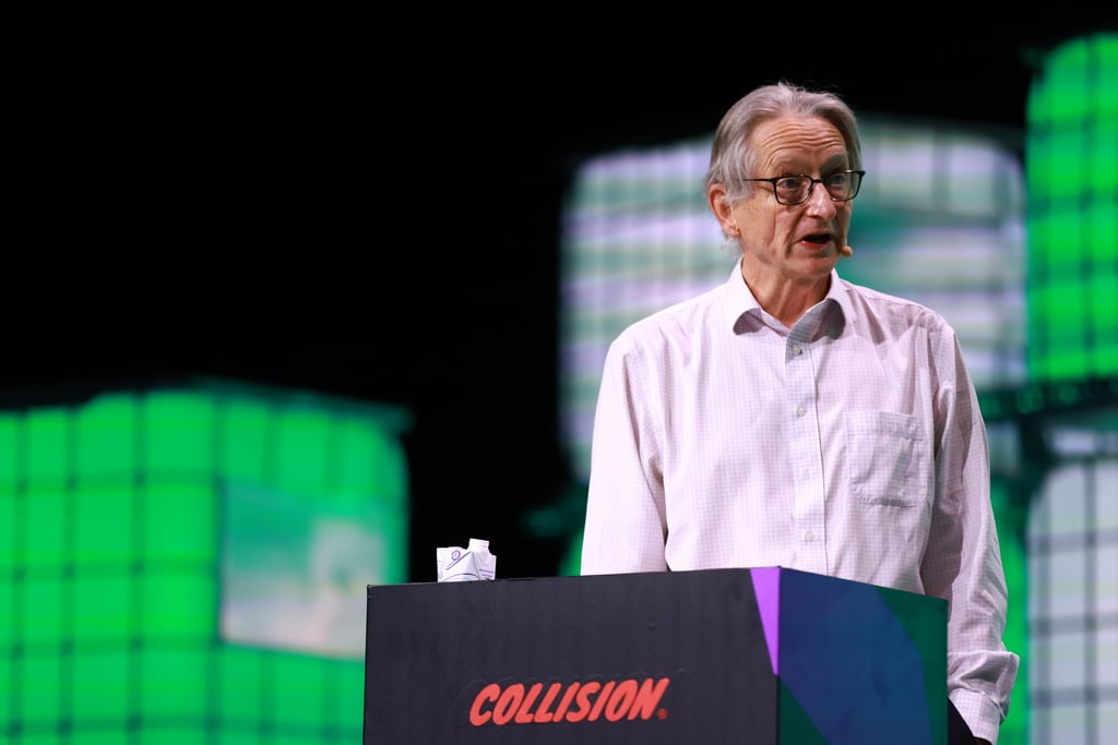 Geoffrey Hinton i hvit skjorte taler fra en talerstol på en scene med grønne lysskjermer bak seg