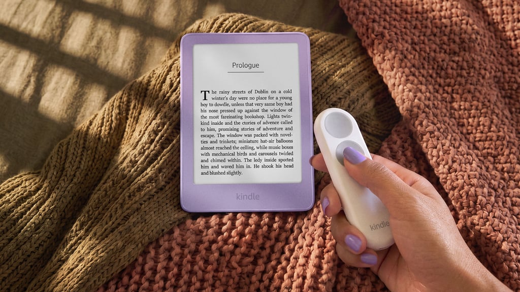 Hånd holder en hvit fjernkontroll ved siden av en lilla Kindle på et strikket pledd