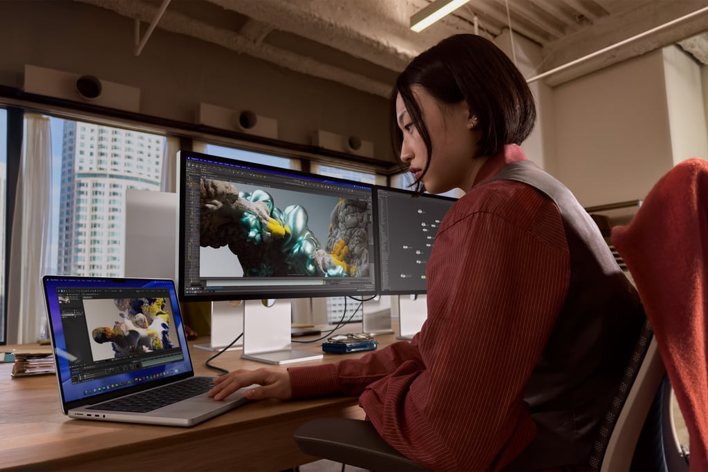 Kvinne ved et skrivebord jobber med 3D-grafikk på en MacBook Pro og to skjermer