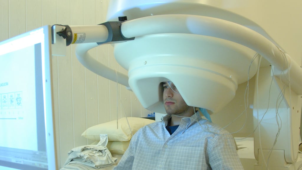 En mann sitter med hodet inne i en stor hvit MEG-maskin og ser på en skjerm