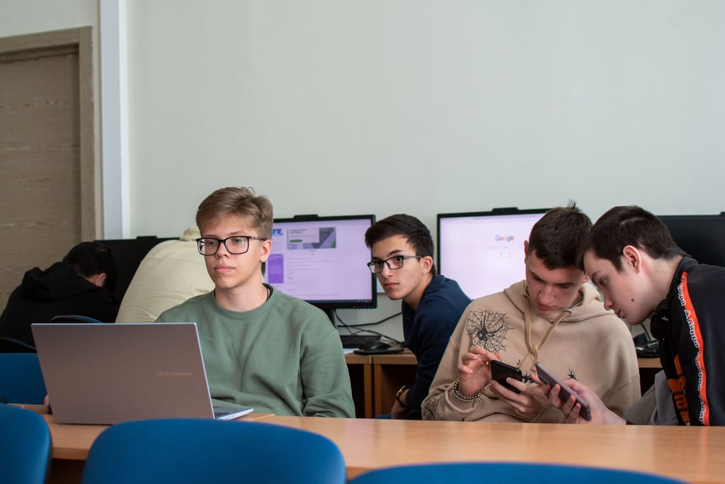 Tenåringer i et klasserom med bærbar PC foran seg og PC-skjermer i bakgrunnen, to av dem ser på mobiltelefoner