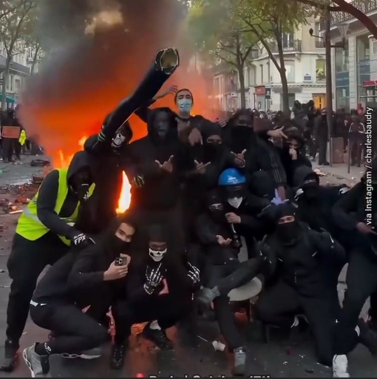 En gruppe maskerte unge menn i svarte klær poserer foran et bål i en gate i Paris