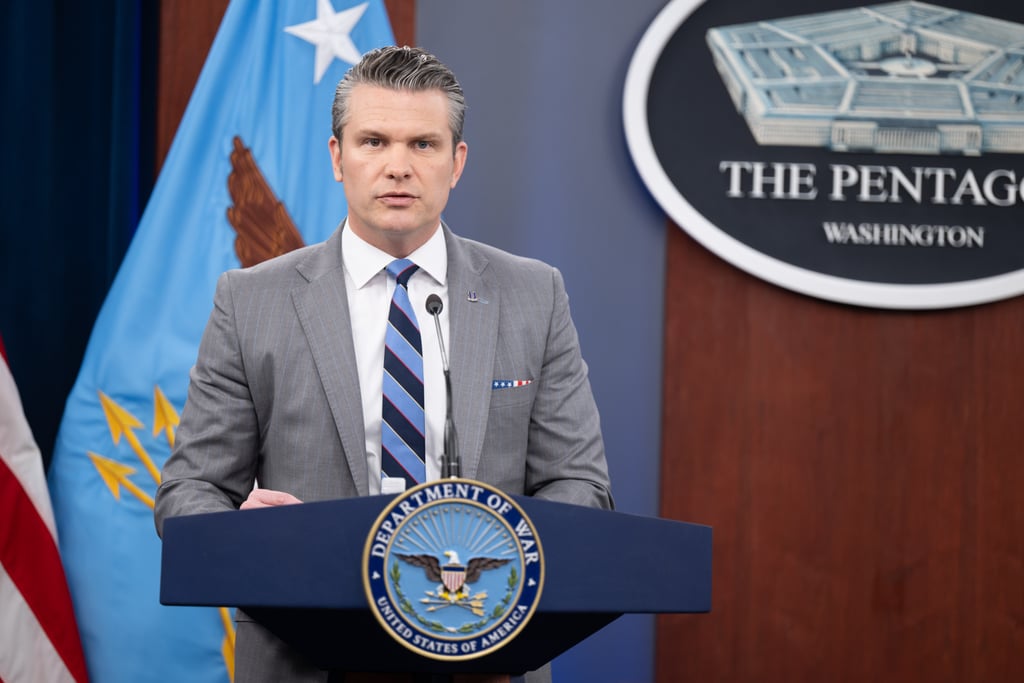 Pete Hegseth i grå dress bak en talerstol med Pentagons emblem i bakgrunnen