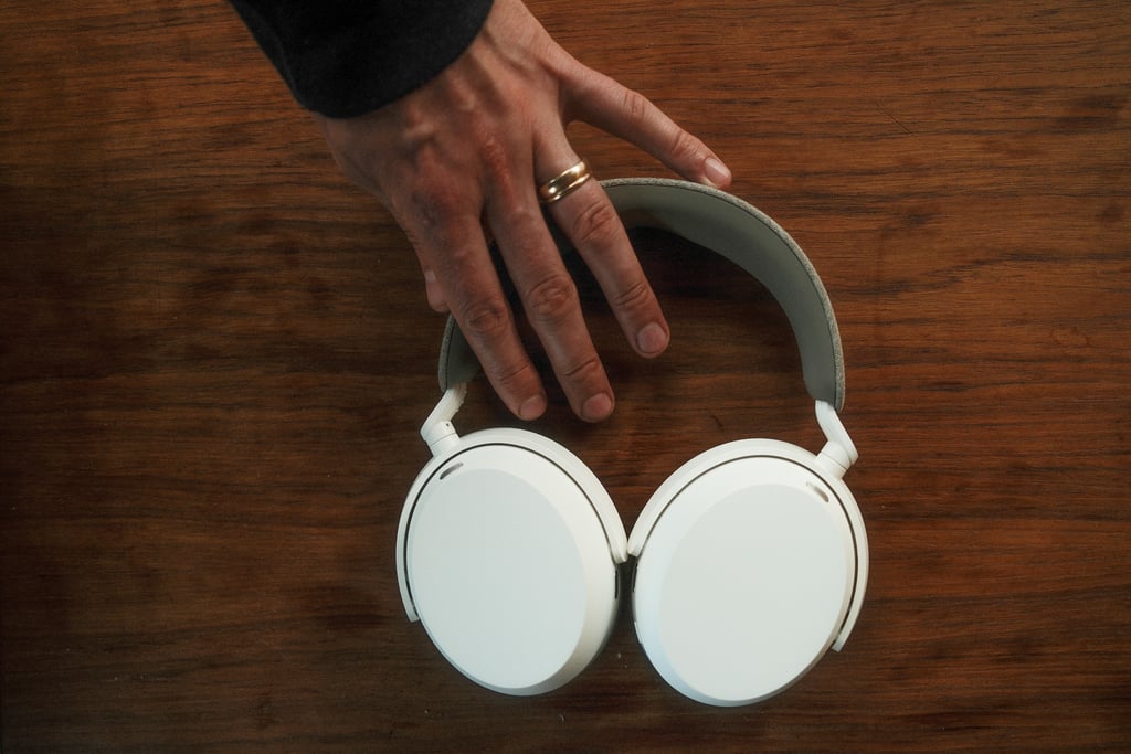 Hånd som løfter Sennheiser Momentum 5 etter bøylen, mot et mørkt trebord