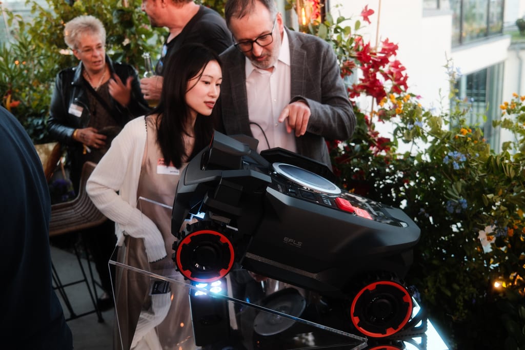 Robotgressklipperen Navimow H5 Pro står på skrå på en gjennomsiktig sokkel mellom blomster på en takterrasse i Berlin, mens to besøkende studerer den