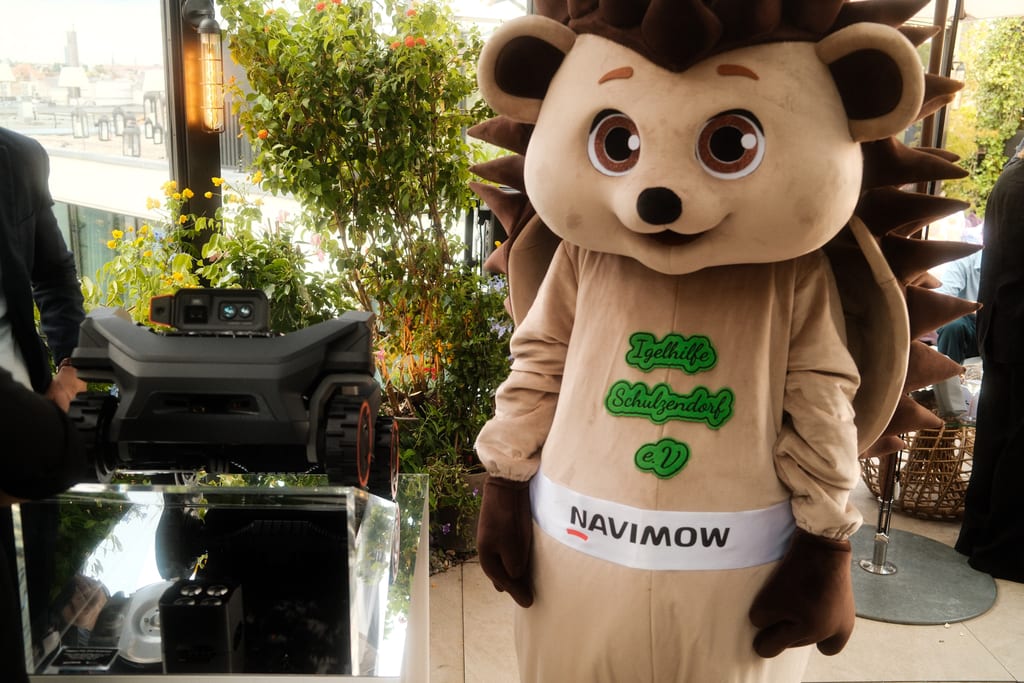Pinnsvinmaskot fra Igelhilfe Schulzendorf med Navimow-banner rundt magen står ved siden av Navimow H5 Pro på en takterrasse