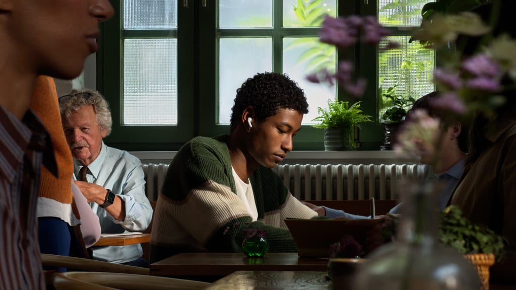 Ung mann med AirPods 5 i øret leser konsentrert på en kafé med folk rundt seg