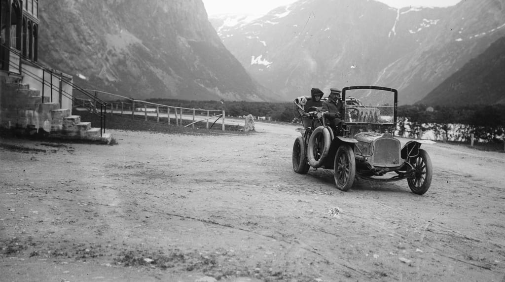 En åpen veteranbil med to personer kjører på en grusplass foran et trehus, med bratte fjell bak. Svart-hvitt foto.