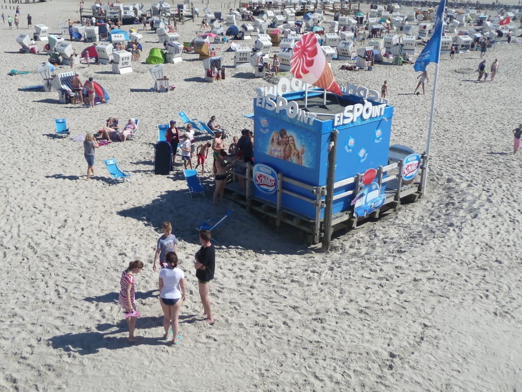Folksom sandstrand sett ovenfra, med en blå iskiosk midt i bildet og badegjester samlet rundt den