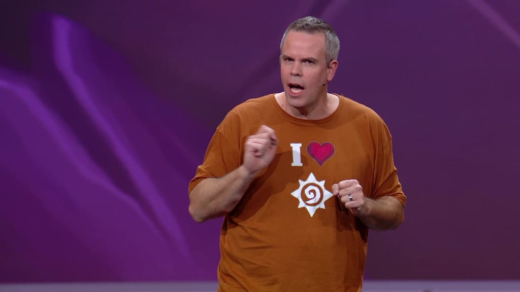 En Hearthstone-utvikler på BlizzCon-scenen i oransje T-skjorte med «I ♥ Hearthstone»-trykk, foran lilla bakgrunn