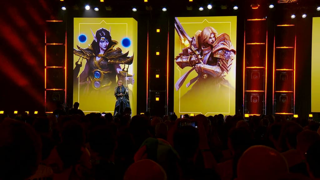 Holly Longdale alene på BlizzCon-scenen i mørk kappe, flankert av to gule storskjermer med Warcraft-figurer, salen full av mobiler i været