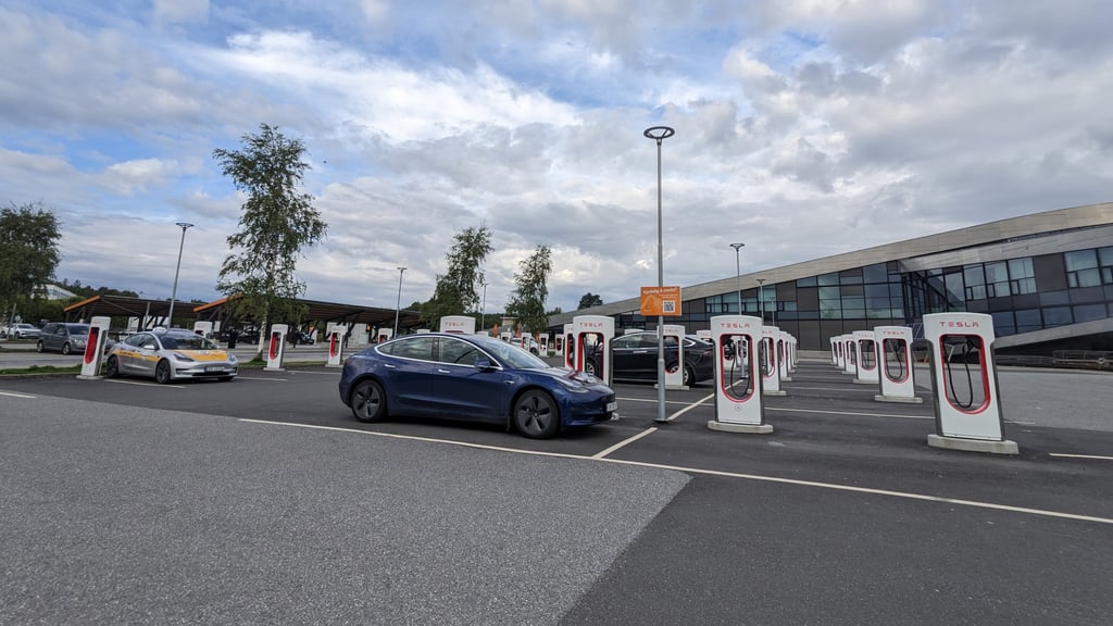 Blå Tesla ved en stor ladestasjon med rader av hvite og røde ladere