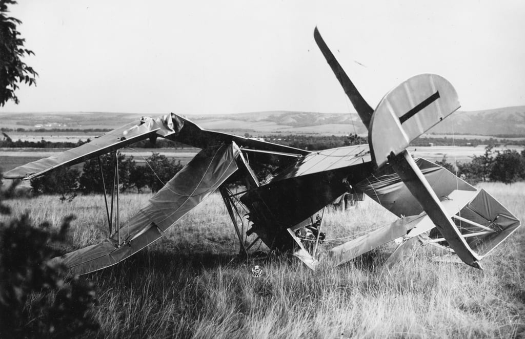 Et havarert dobbeltdekkerfly ligger med nesa i bakken på et jorde. Svart-hvitt foto.
