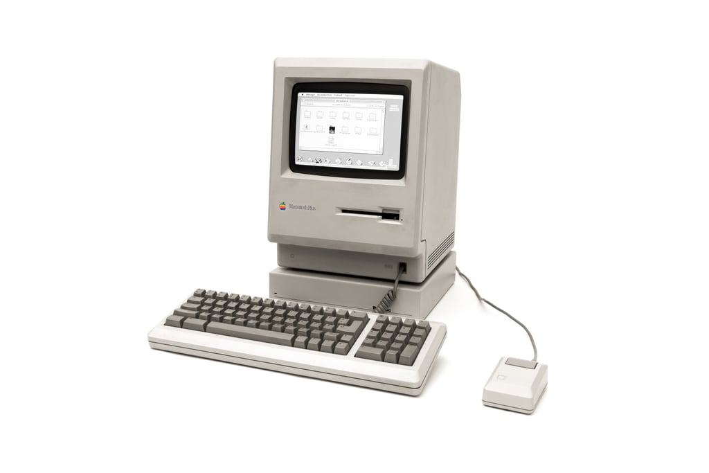 En beige Macintosh Plus med tastatur og mus mot hvit bakgrunn. Skjermen viser et vindu med mapper og en programlinje der Excel er ett av programmene.
