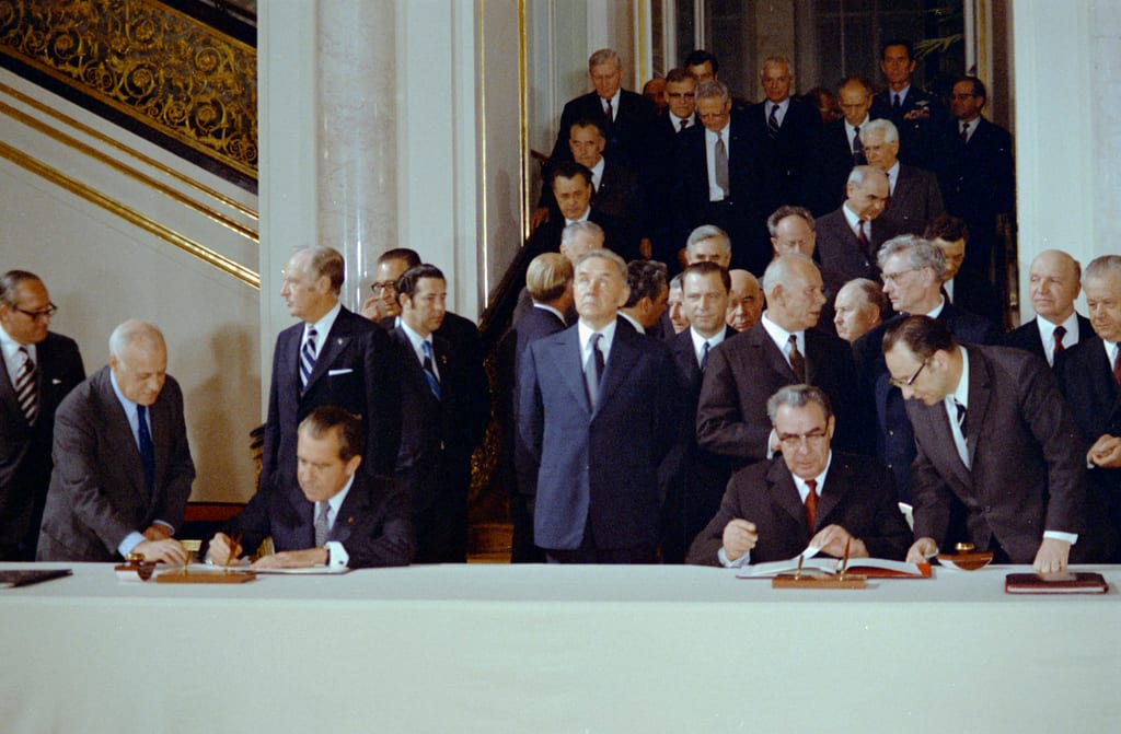 Richard Nixon og Leonid Bresjnev sitter ved et langt bord og signerer dokumenter, med rader av menn i dress bak seg.
