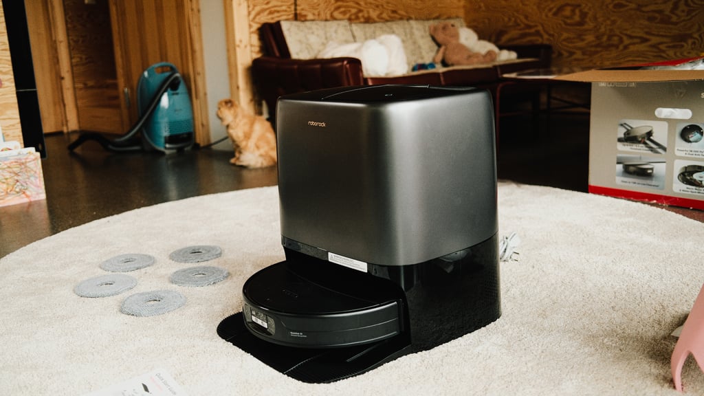 Roborock Saros 20 Neo i dokken på et rundt teppe, med fem moppeputer, en pappeske og en katt i bakgrunnen