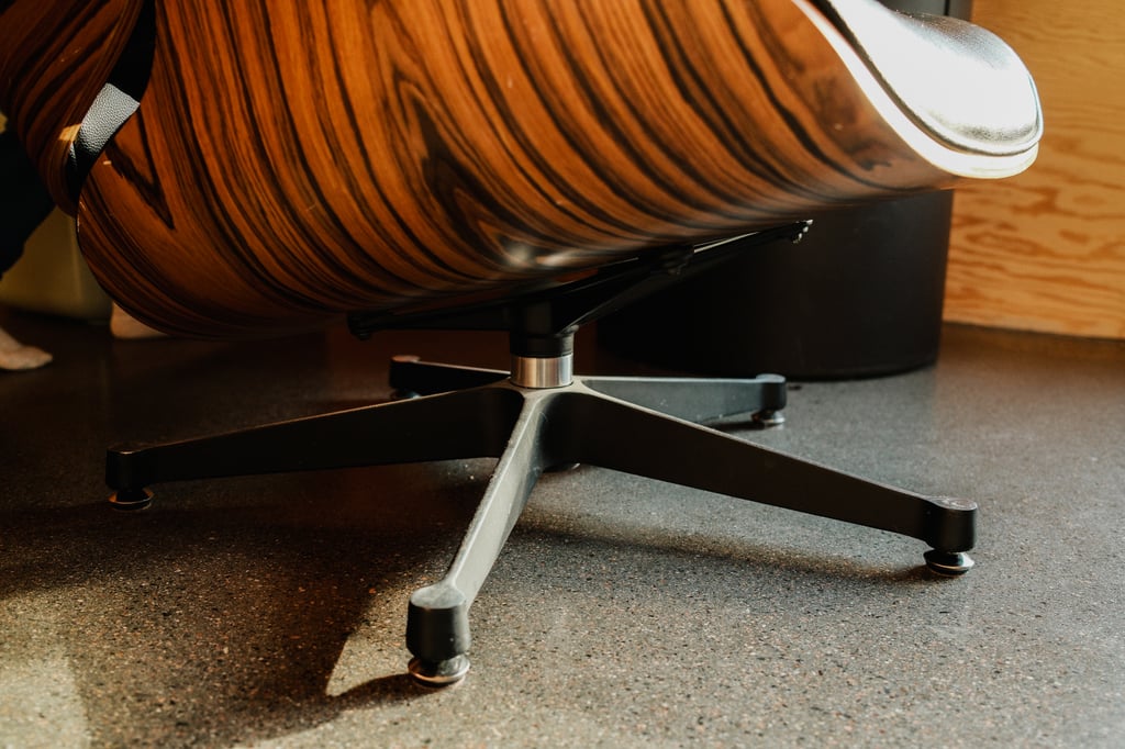 Foten under en Eames-stol av tre og svart skinn, sett fra gulvnivå i sollys