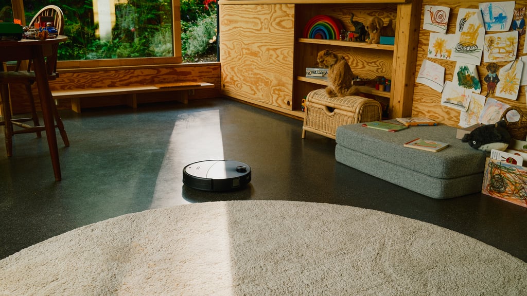 Roborock Saros 20 Neo på stuegulvet i sollys, med en perserkatt på en kurvkiste og et rundt teppe i forgrunnen