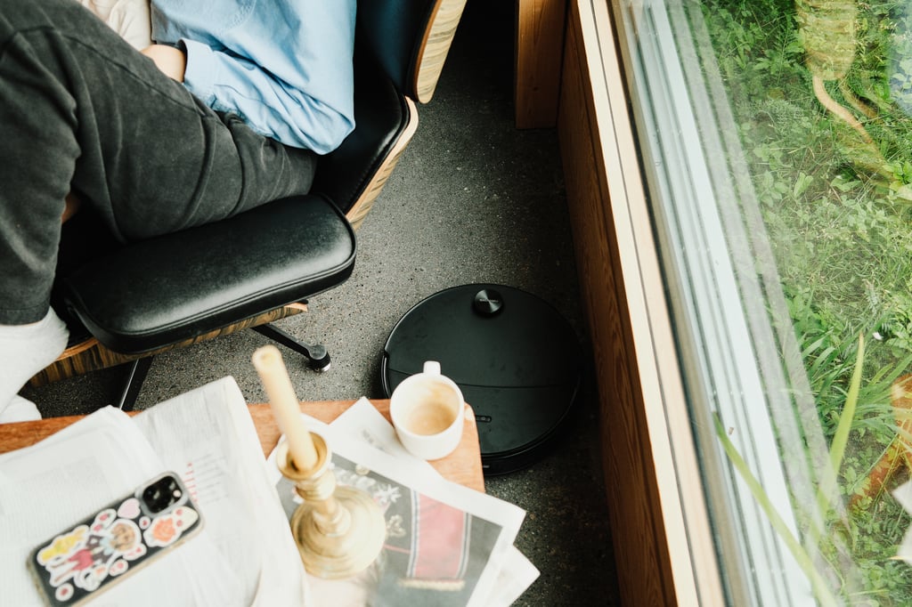 Roborock Saros 20 Neo ved et vindu, ved siden av en person i Eames-stol og et bord med kaffekopp