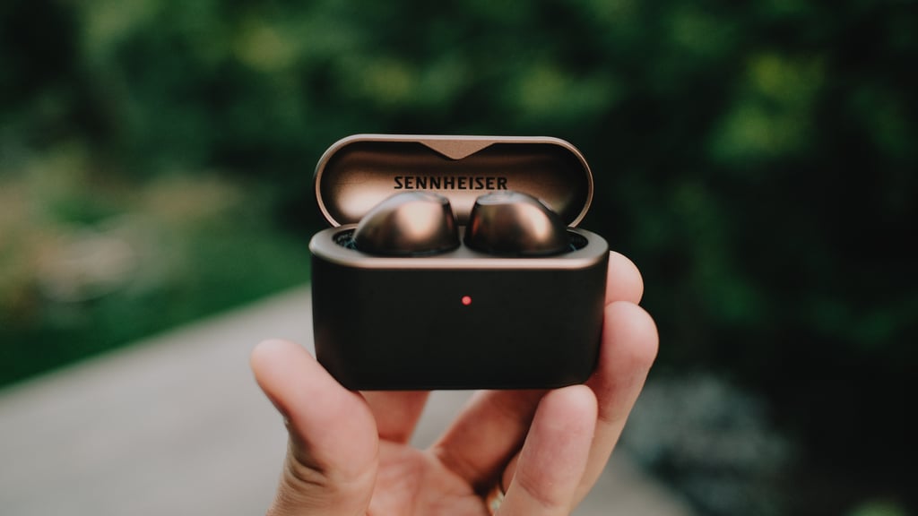 Åpent Sennheiser Momentum True Wireless 5-etui i en hånd, proppene i kobber mot grønt