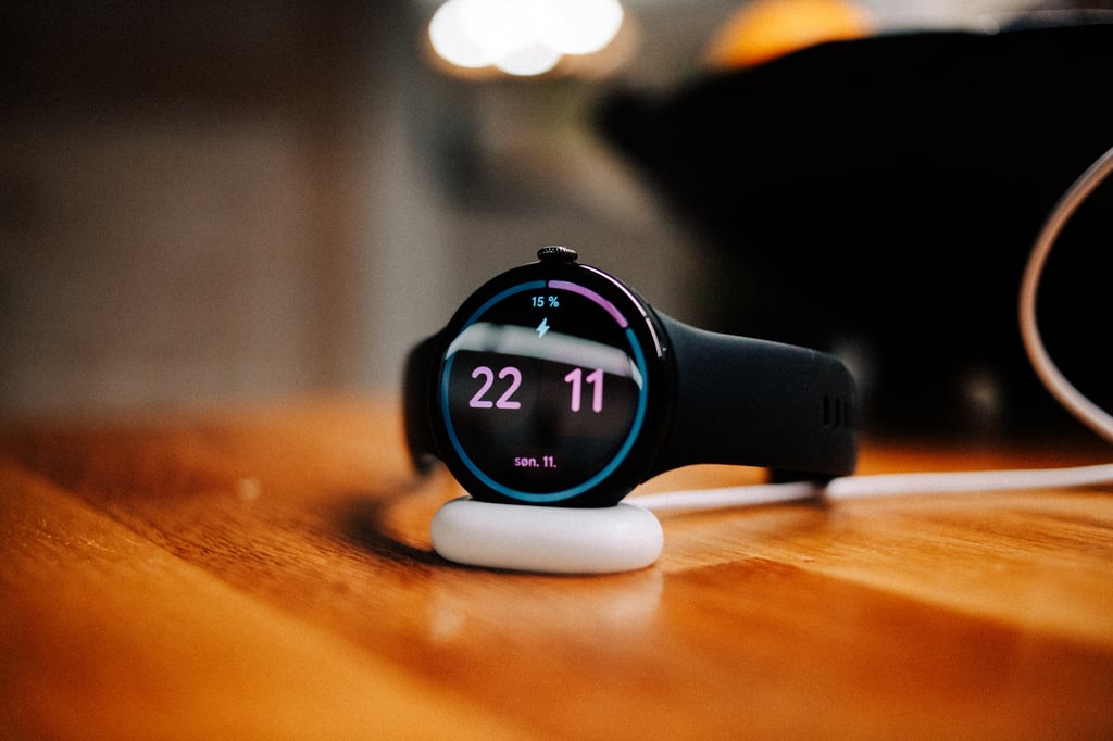 Pixel Watch 5 står på den hvite laderen på et trebord og viser 15 prosent batteri