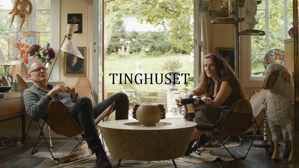 En mann og en kvinne sitter i lenestoler i en lys stue full av kunst og antikviteter